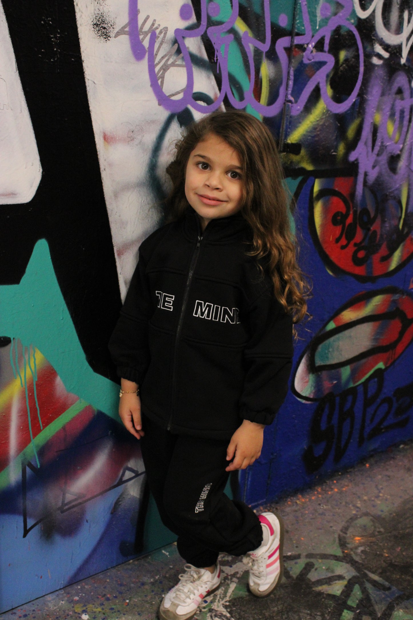 Smiling young girl in black 'THE MIND' tracksuit set with white and pink sneakers, standing against a vibrant graffiti wall.
