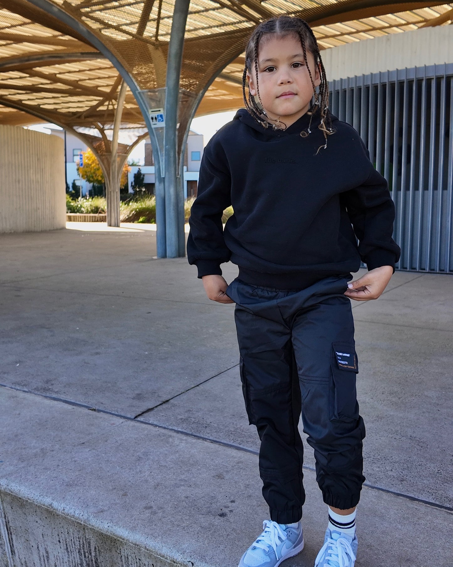 Young child wearing a black "the minis" hooded sweatshirt and black cargo track pants with side pockets.