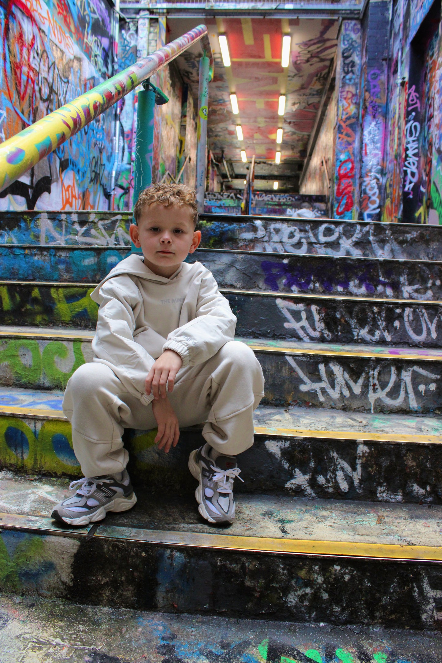 Kid's off-white 'The Minis' tracksuit and gray sneakers, worn by a boy sitting on colorful graffiti stairs.