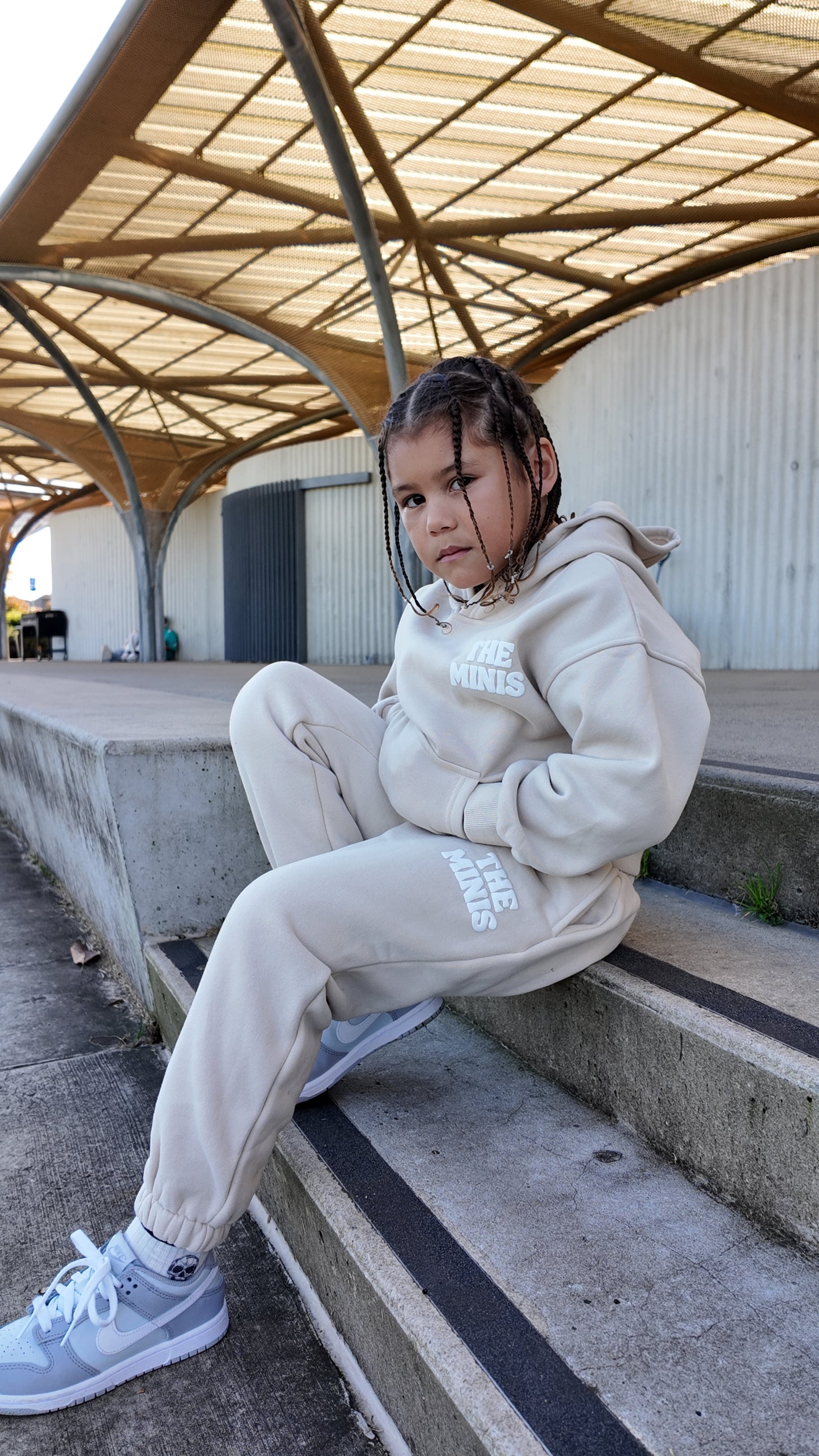 Kid wearing a cream 'THE MINIS' tracksuit with a hoodie and matching joggers, sitting on concrete steps.