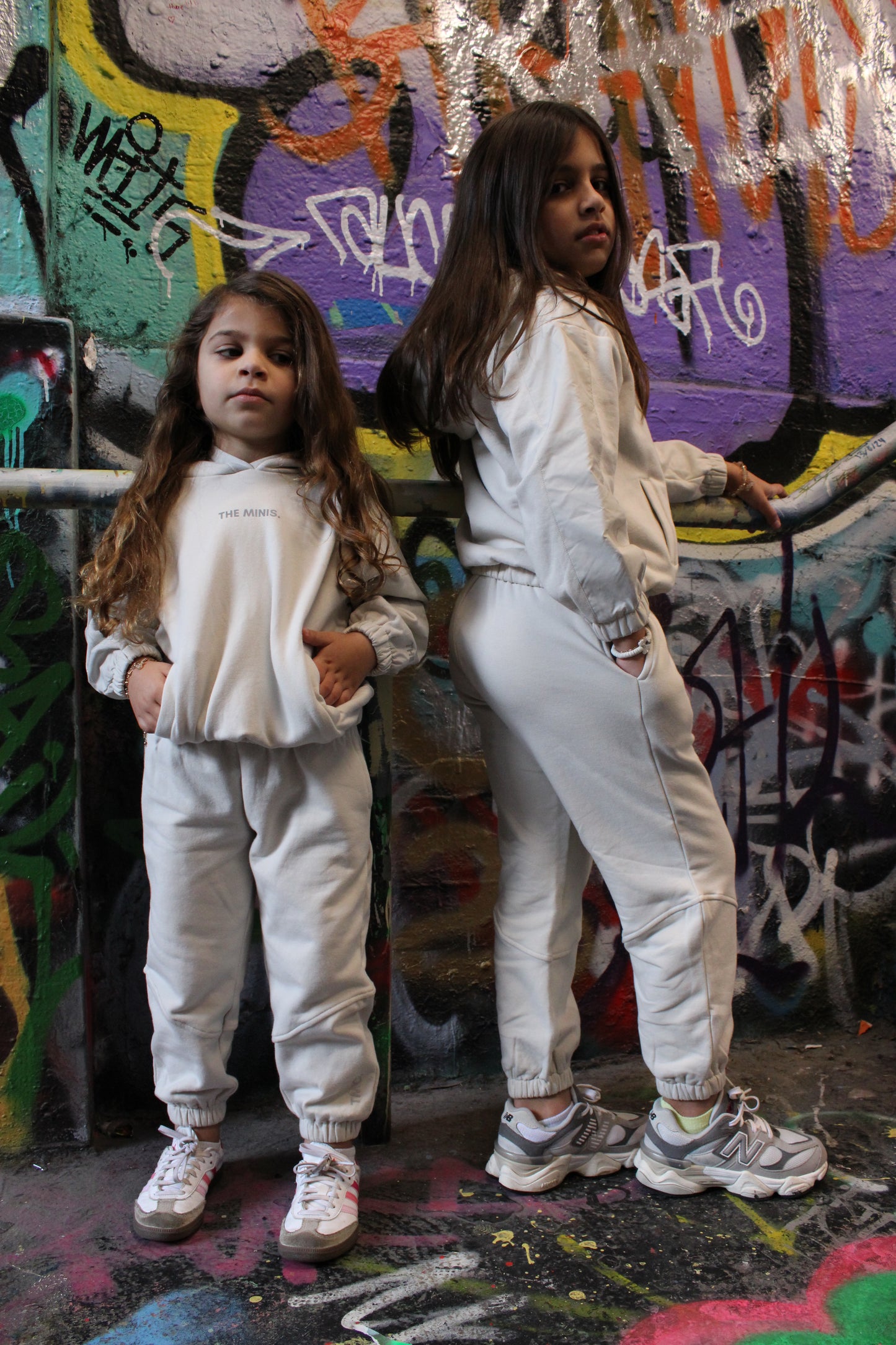 Two girls in matching off-white 'The Minis' hoodies and joggers standing against a vibrant graffiti wall.
