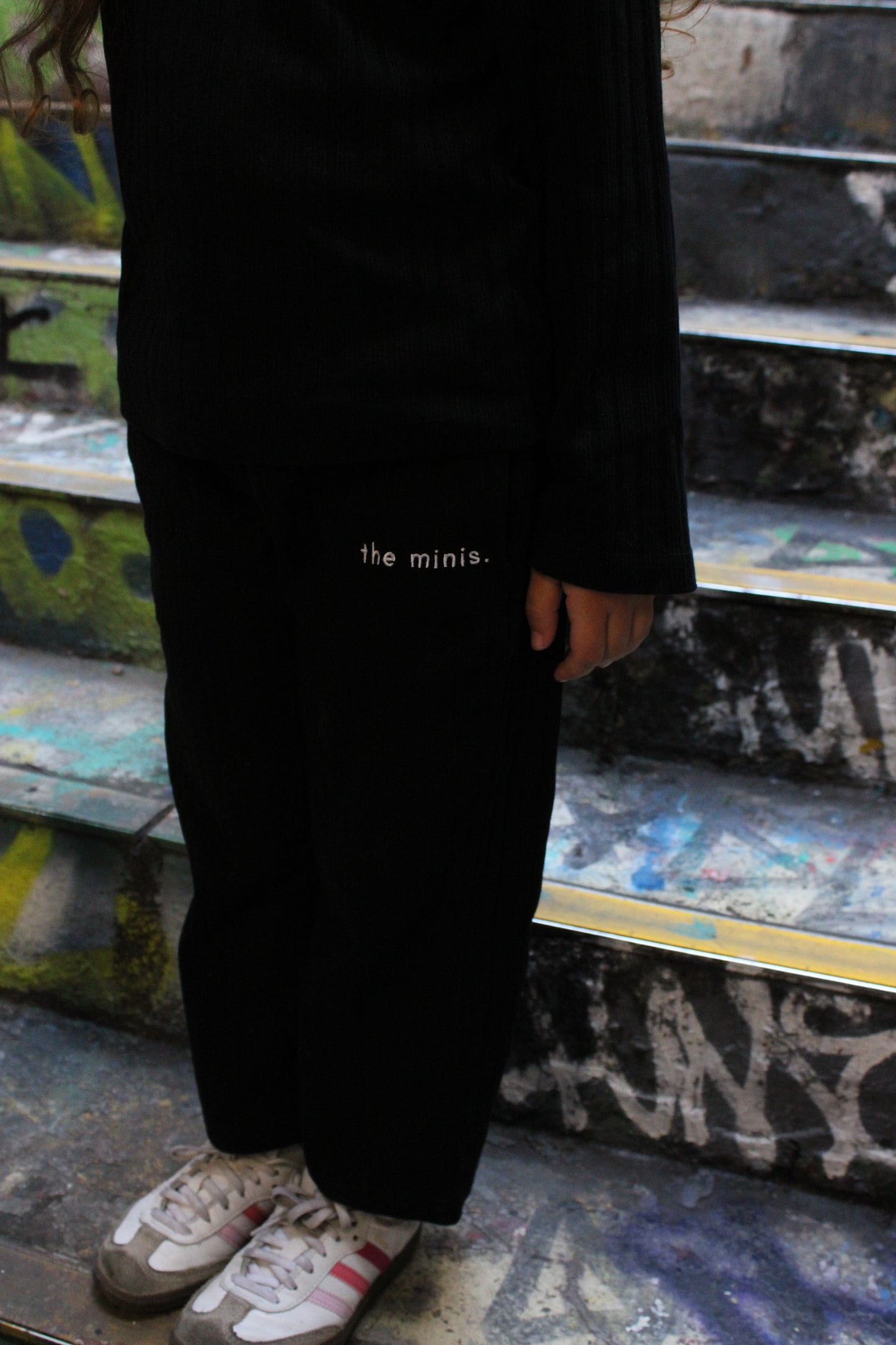 Black pants with 'the minis.' text, black long-sleeve top, and sneakers worn by a person on graffiti steps.