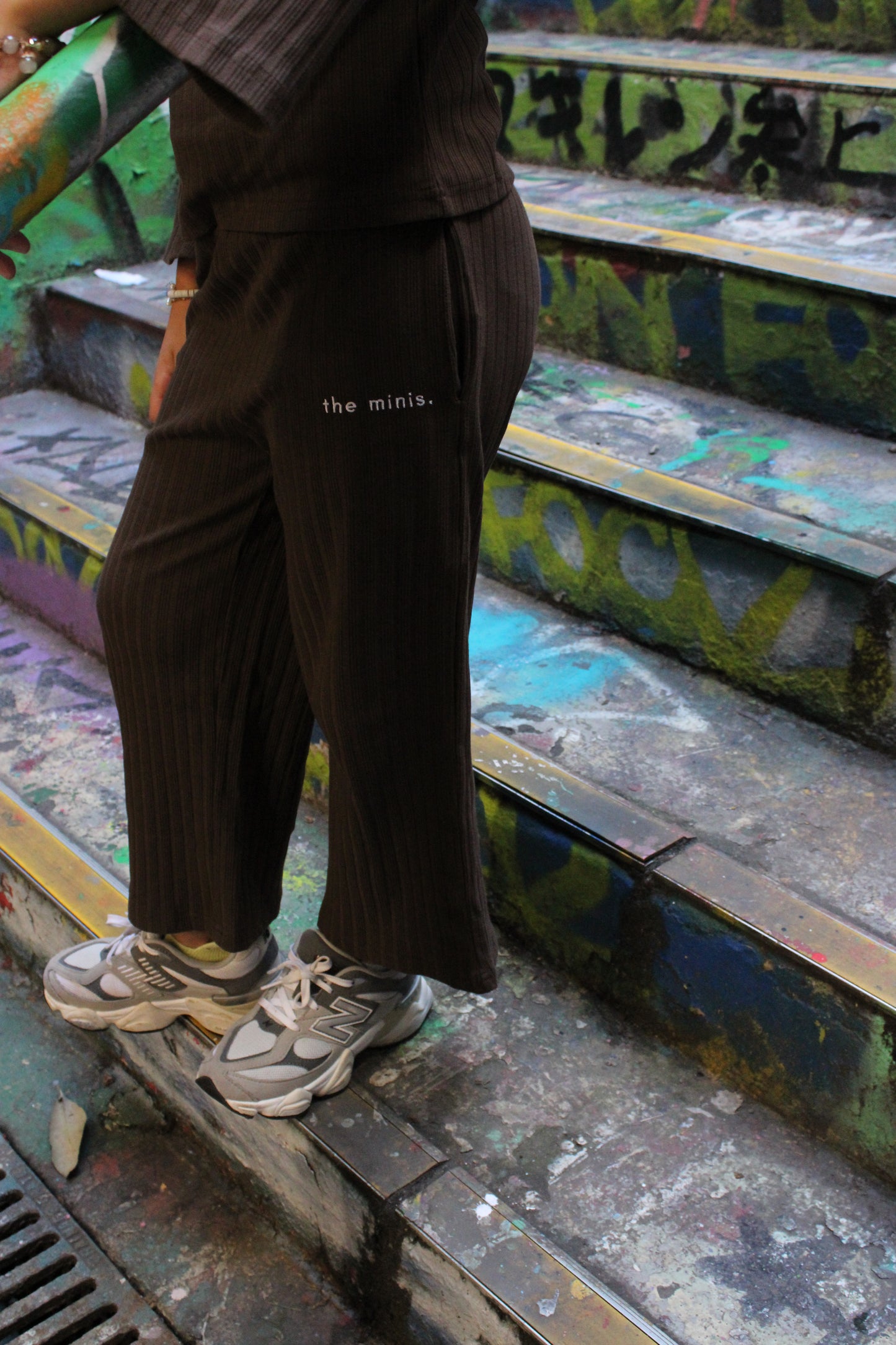 Person wearing brown ribbed wide-leg pants with 'the minis.' logo and grey New Balance sneakers on graffiti-covered stairs.
