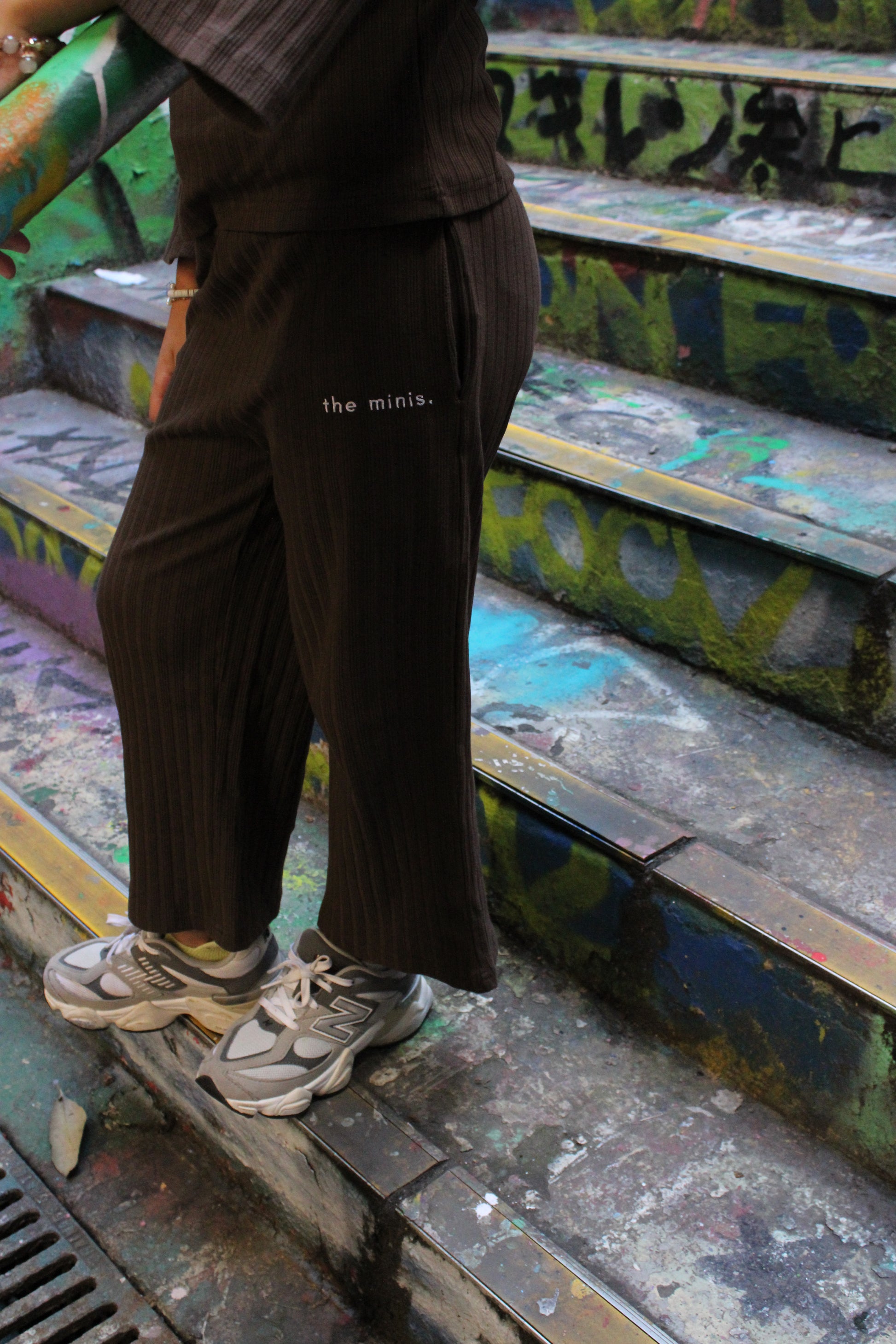 Person wearing brown ribbed wide-leg pants with 'the minis.' logo and grey New Balance sneakers on graffiti-covered stairs.