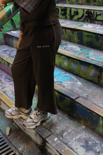 Person wearing brown ribbed wide-leg pants with 'the minis.' logo and grey New Balance sneakers on graffiti-covered stairs.