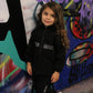 Smiling young girl in black 'THE MIND' tracksuit set with white and pink sneakers, standing against a vibrant graffiti wall.