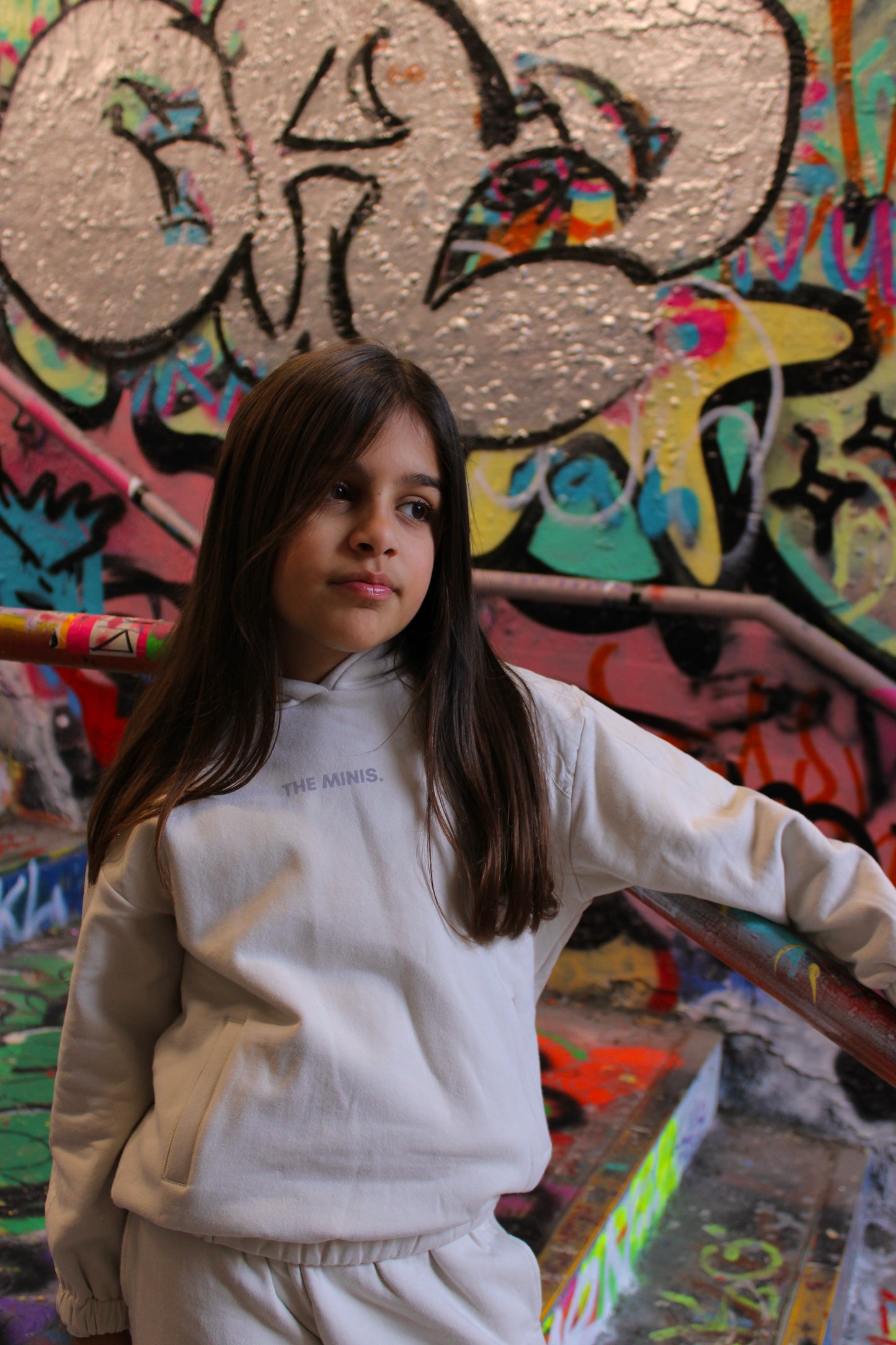 Child in white hoodie and pants with 'THE MINIS.' text, standing against a vibrant graffiti backdrop.