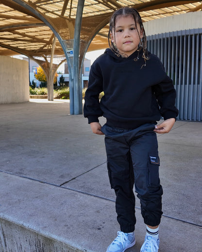 Young child wearing a black "the minis" hooded sweatshirt and black cargo track pants with side pockets.