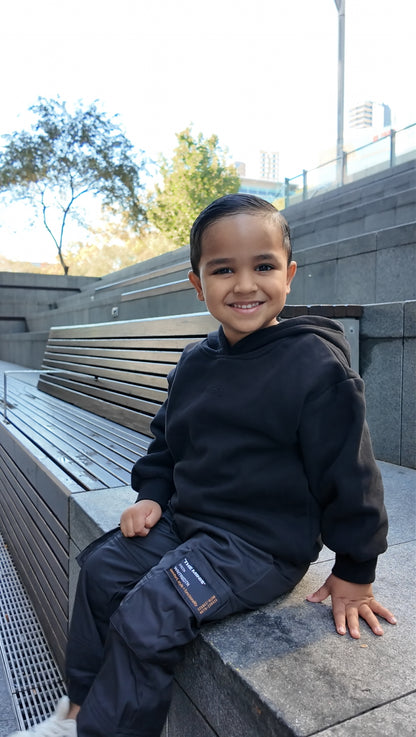 Smiling young boy wearing "THE MINIS" black hoodie & "WNTREDTN" black cargo pants with "comfort, style + functionality" label.