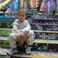 Kid's off-white 'The Minis' tracksuit and gray sneakers, worn by a boy sitting on colorful graffiti stairs.