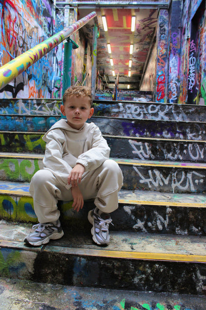 Kid's off-white 'The Minis' tracksuit and gray sneakers, worn by a boy sitting on colorful graffiti stairs.