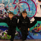 Kids black "THE MINIS" tracksuit on two young boys against a vibrant graffiti wall.