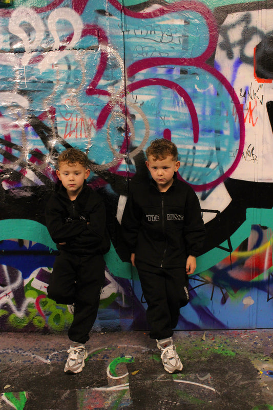 Kids black "THE MINIS" tracksuit on two young boys against a vibrant graffiti wall.