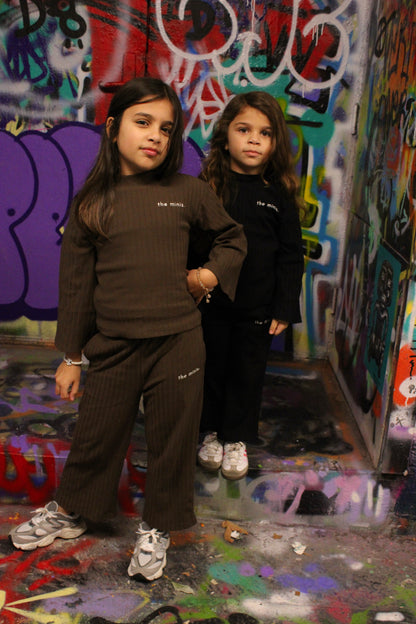 Two girls wearing 'the minis.' brown and black ribbed loungewear sets, featuring long-sleeve tops and matching pants.