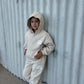 Young child wearing a beige "The Minis" hoodie and jogger tracksuit set, leaning against a textured wall.