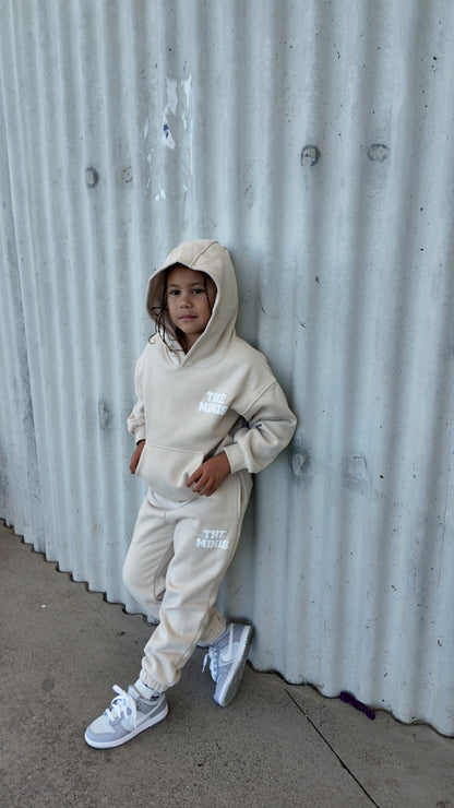 Young child wearing a beige "The Minis" hoodie and jogger tracksuit set, leaning against a textured wall.