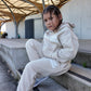 Kid wearing a cream 'THE MINIS' tracksuit with a hoodie and matching joggers, sitting on concrete steps.