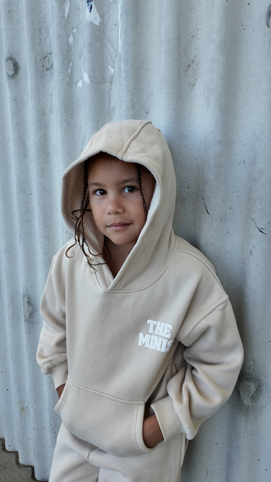 Young child wearing a beige 'THE MINIS' hoodie, with braids, looking at the camera.