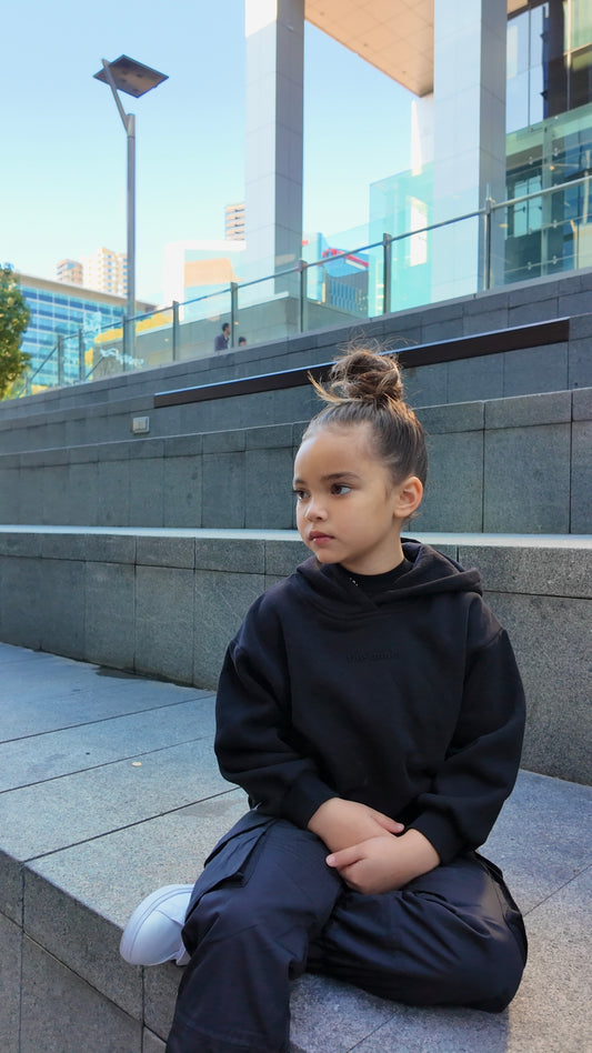 Young child wearing a black pullover hoodie, dark cargo pants, and white sneakers, sitting on concrete steps.