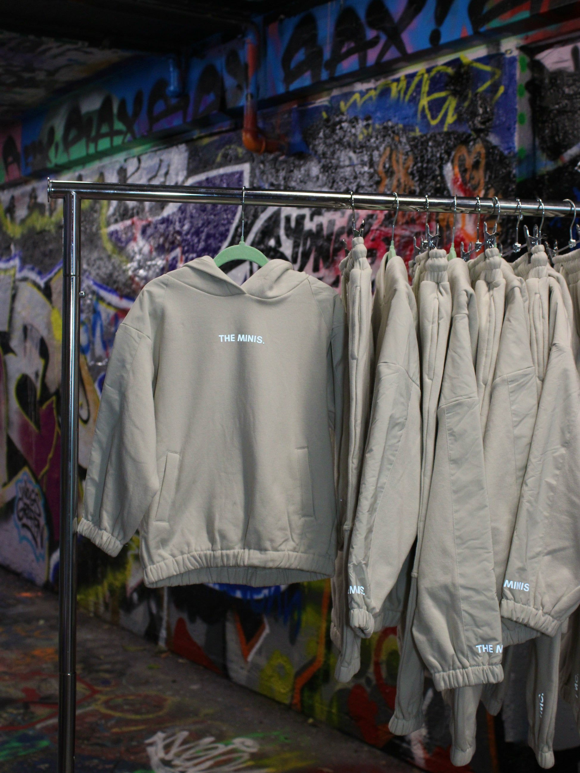 Tan "THE MINIS." hoodie and matching sweatpants set on a rack with graffiti background