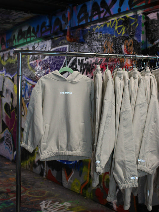 Tan "THE MINIS." hoodie and matching sweatpants set on a rack with graffiti background