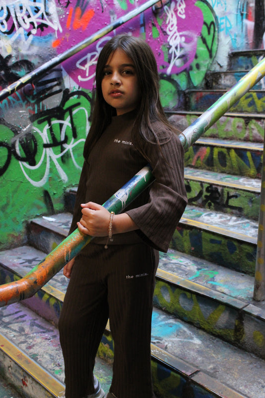 Child wearing brown ribbed "the minis." long-sleeve top and matching pants, standing on graffiti-covered stairs.