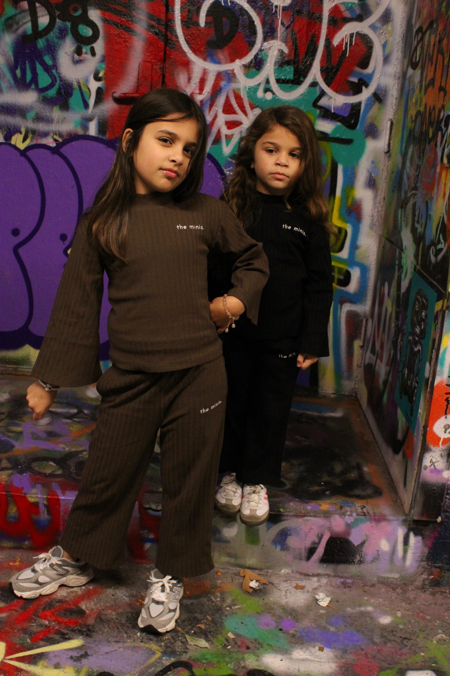 Kids wearing 'the minis.' ribbed brown and black long-sleeve outfits, street art background.