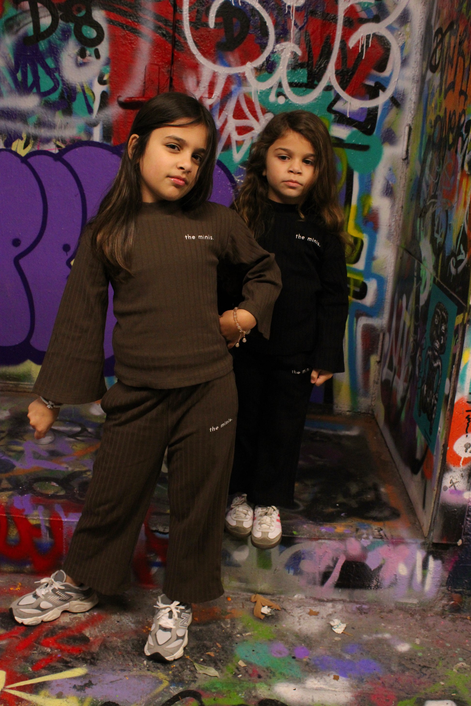 Kids wearing 'the minis.' ribbed brown and black long-sleeve outfits, street art background.