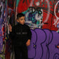 Young boy wearing a black "THE MINIS" tracksuit and white sneakers, standing against a colorful graffiti wall.