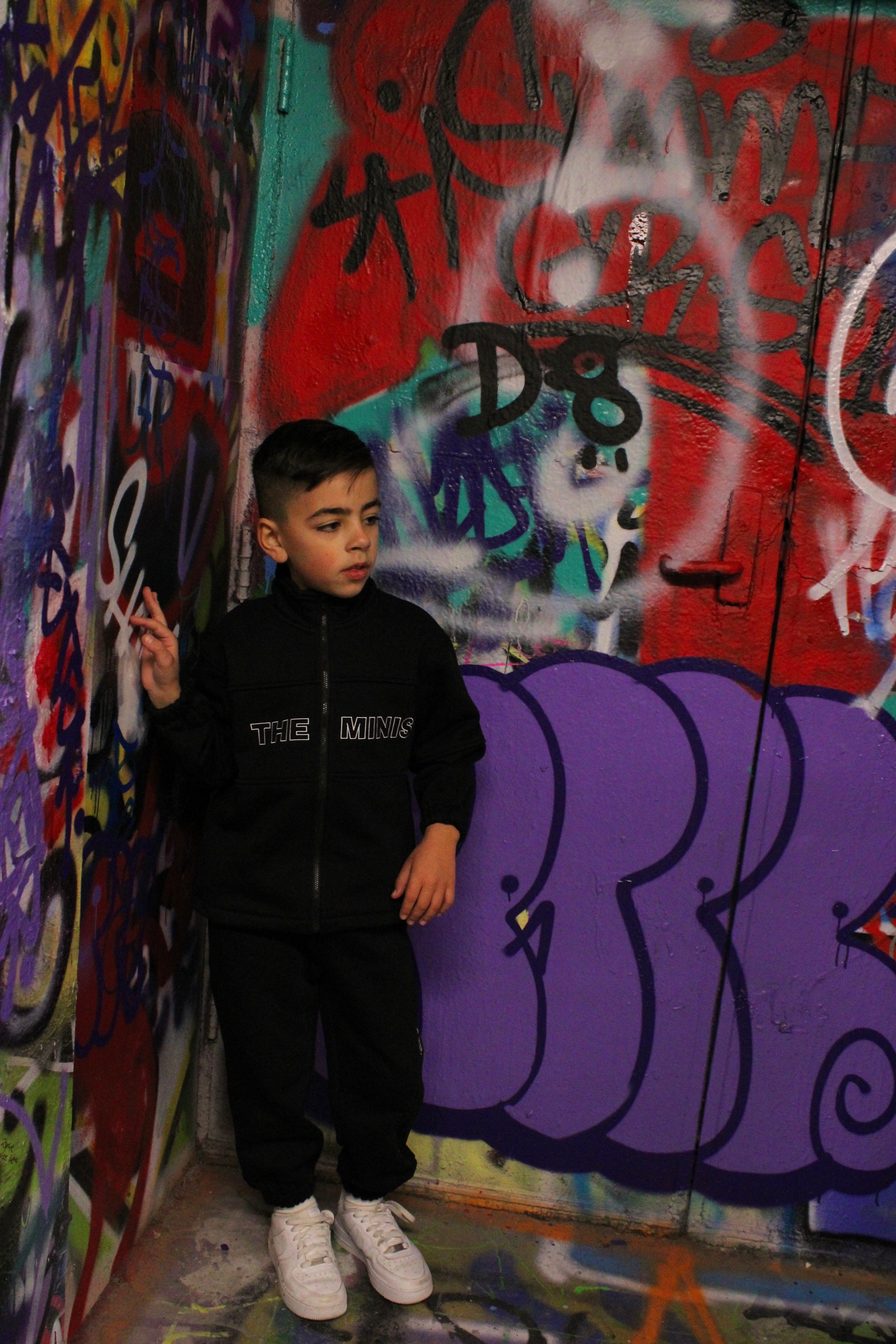 Young boy wearing a black "THE MINIS" tracksuit and white sneakers, standing against a colorful graffiti wall.