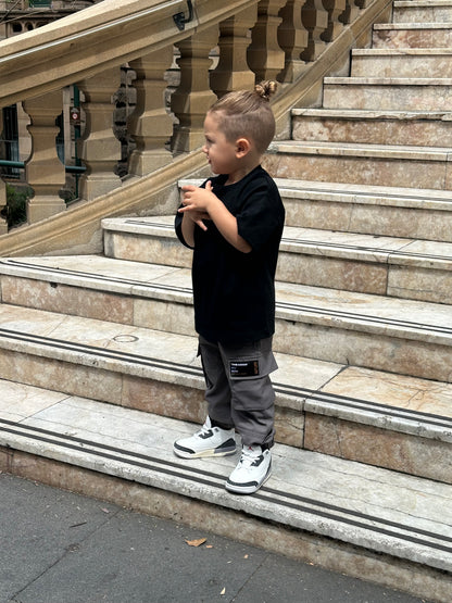 Young boy wearing black t-shirt, gray 'THE MINIS' cargo pants, and white sneakers.