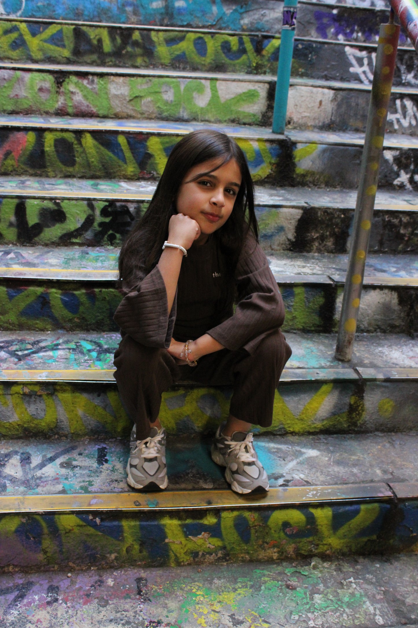 Young girl wearing a brown ribbed long-sleeve top & matching pants with grey athletic shoes, sitting on graffiti steps.