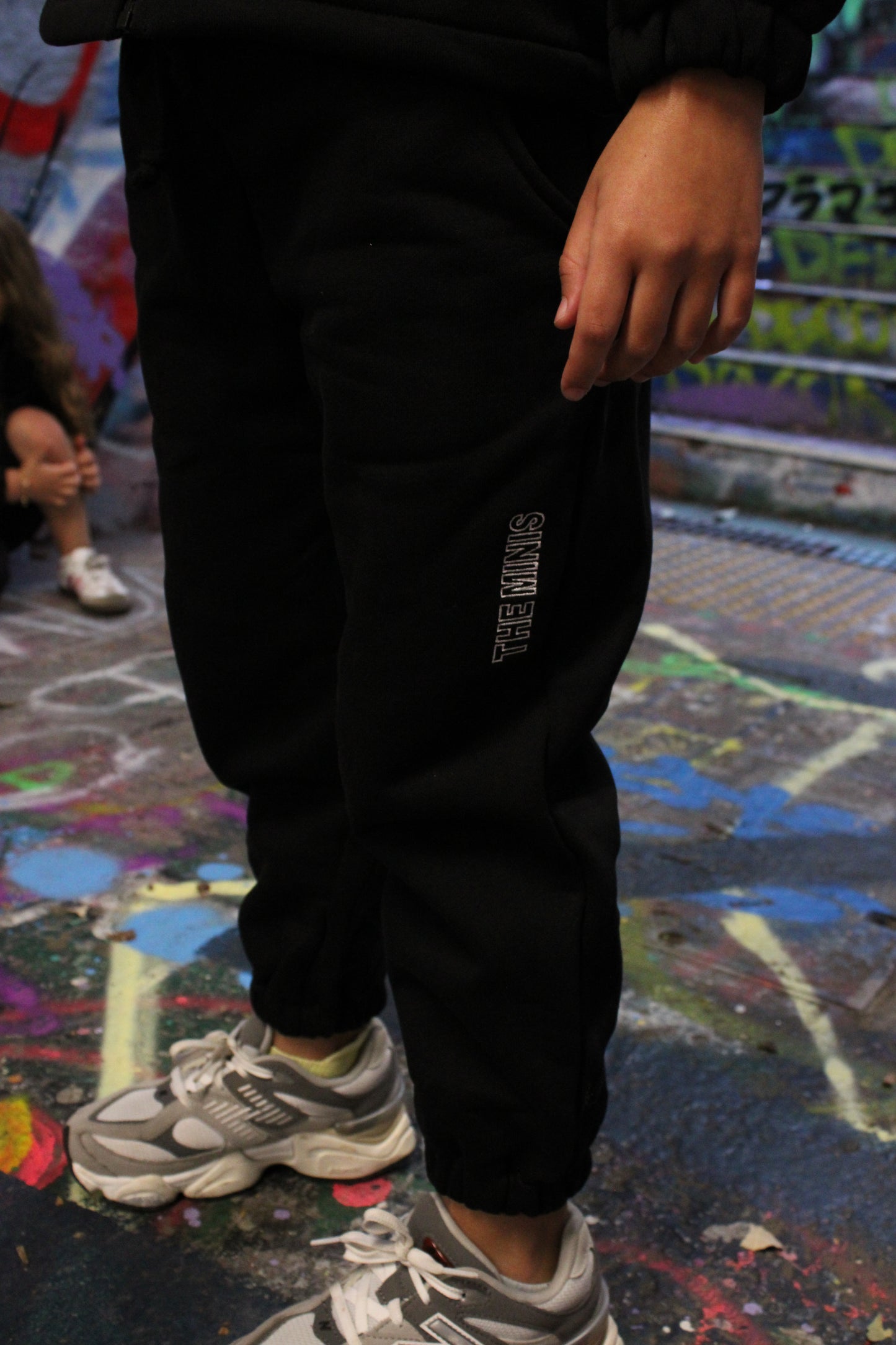 Black sweatpants featuring 'THE MINIS' logo on the side, paired with gray and white sneakers on a graffiti-covered surface.
