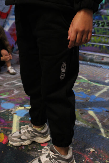 Black sweatpants featuring 'THE MINIS' logo on the side, paired with gray and white sneakers on a graffiti-covered surface.
