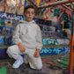 A young boy in a beige hoodie and joggers poses on colorful graffiti-covered steps, wearing white sneakers.