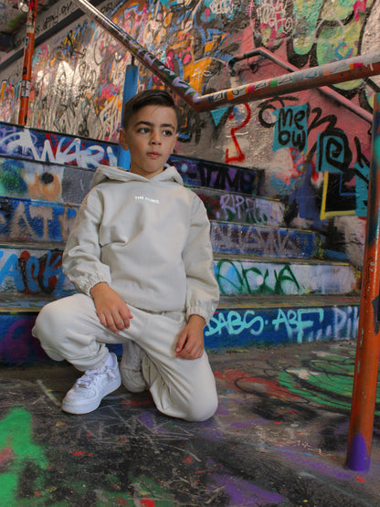 A young boy in a beige hoodie and joggers poses on colorful graffiti-covered steps, wearing white sneakers.