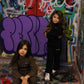 Two young girls wearing 'the minis' matching loungewear sets, one in black and one in brown, posed against a vibrant graffiti-covered wall.