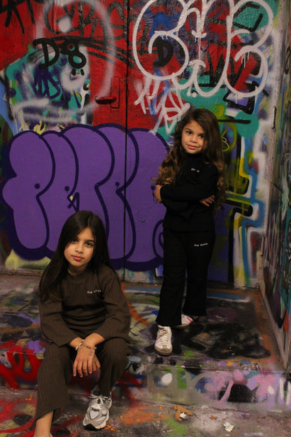 Two young girls wearing 'the minis' matching loungewear sets, one in black and one in brown, posed against a vibrant graffiti-covered wall.