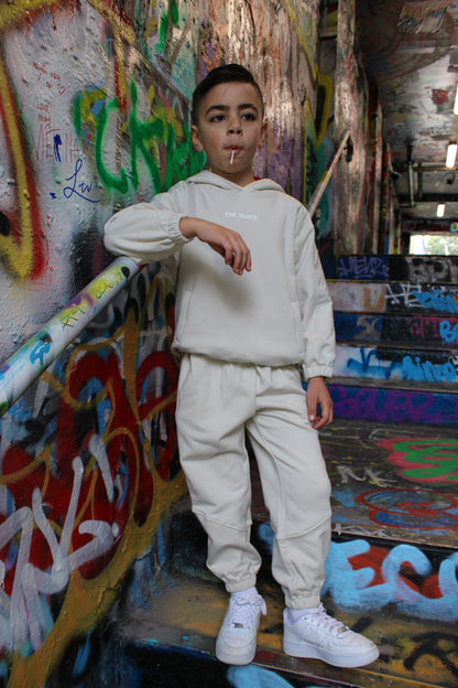 Young boy wearing a cream 'THE MINIS.' kids' tracksuit, including a hoodie and matching joggers, with white sneakers on graffiti stairs.