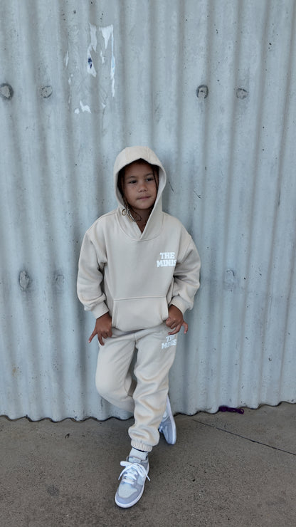 Child wearing a beige The Minis hoodie and matching sweatpants set, standing casually against a wall.