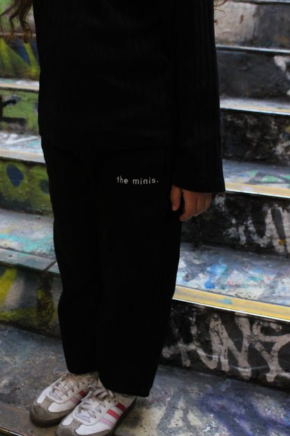 Black pants with 'the minis.' text, black long-sleeve top, and sneakers worn by a person on graffiti steps.
